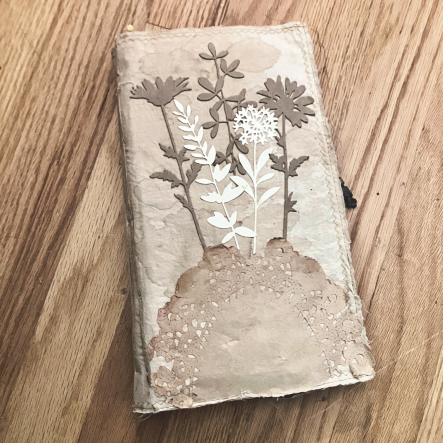 Cream colored muslin journal cover with die cut flowers in shades of brown and white, with a tea dyed doily and machine stitching around the edges.