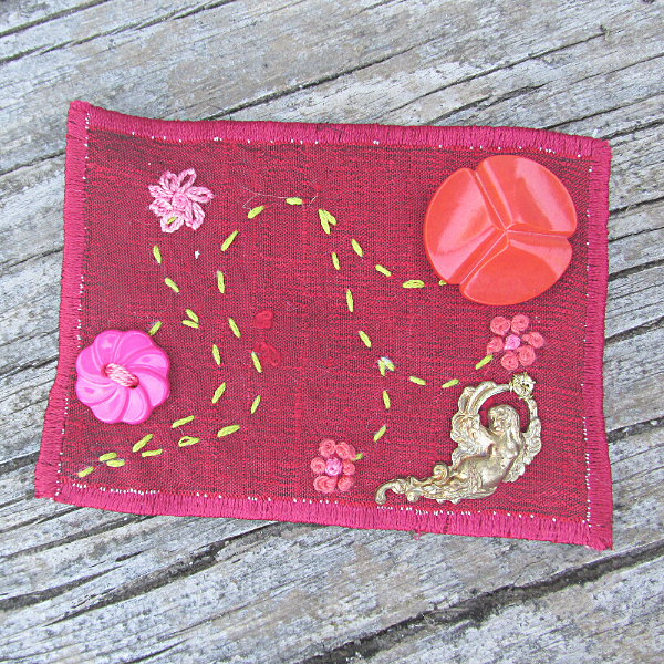 red silk ACEO with embroidered flowers, vintage buttons, and a brass fairy charm