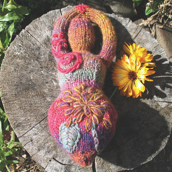 knittted goddess in shades of pink, orange, and green, with embroidered flowers and spirals