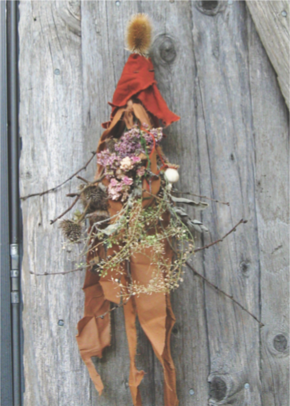spirit doll with a teasel seedpod for a head, wearing orange and brown rags, adorned with dried flowers and seed pods