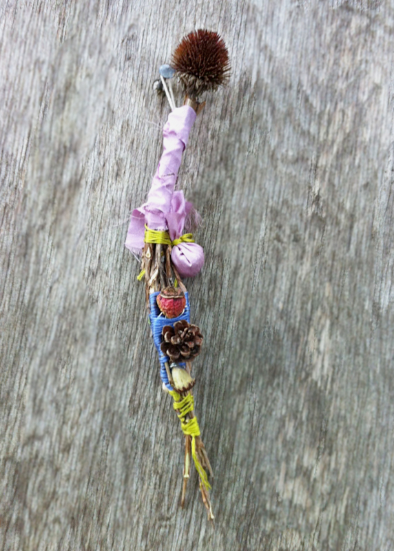 long, thin spirit doll with an echinacea cone for a head, wrapped in colorful cloth and string, adorned with a rose hip and a pine cone