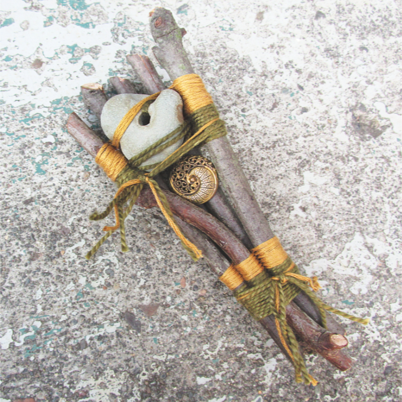 5 twigs woven together with green and yellow thread, adorned with a hagstone and an ornate vintage button