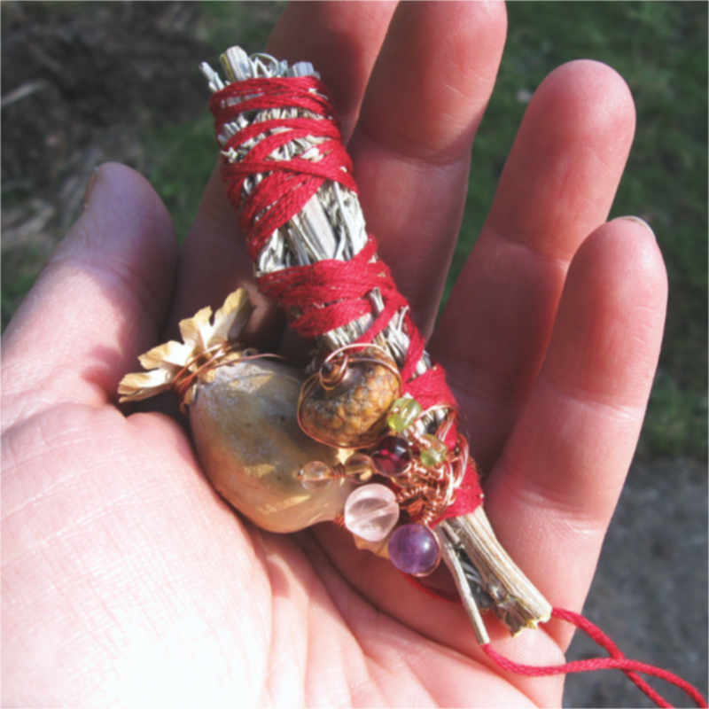 bundle of dried garden sage wrapped with red thread, adorned with a white poppy pod, amethyst, rose quartz, peridot, and a small acorn