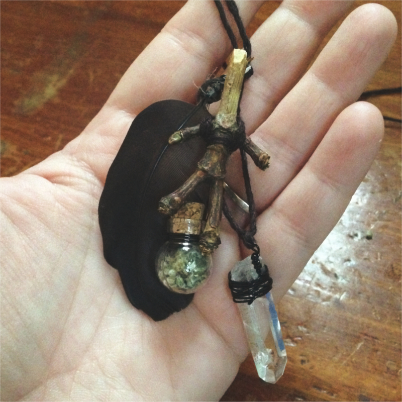 a black feather, quartz point, tiny round jar of mugwort, and an anthropomorphic twig hanging from black thread