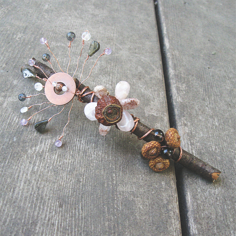 dark brown twig adorned with copper wire, beads, a rose hip, and tiny acorns