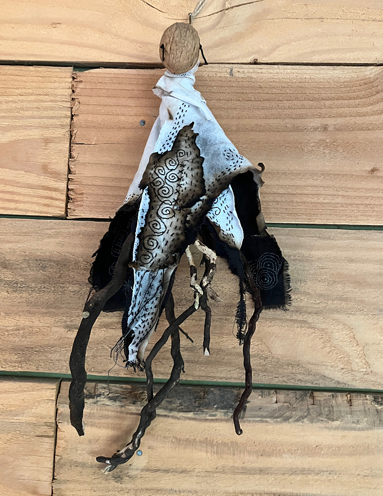 a doll with a walnut shell for a face, gnarly roots for a body, and a tattered, embroidered cloak in black, white, and gray hangs on a wooden wall.