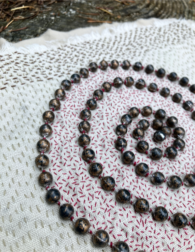 Closeup on a spiral of mottled bronze beads stitched to neutral fabric, surrounded with red seed stitch and lines of running stitch in two shades of brown.