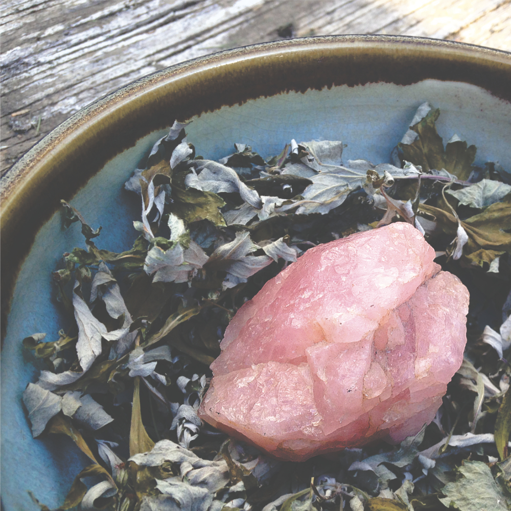 a pottery bowl filled with dried herbs and a piece of rose quartz