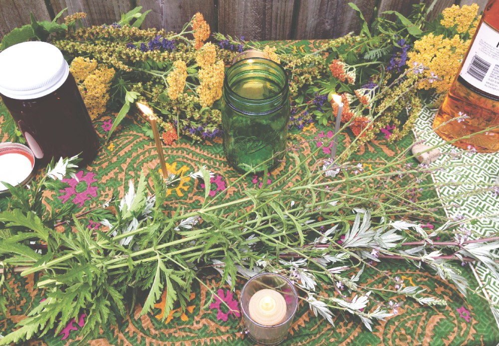 a table covered with herbs, candles, liquor, honey, stones, and a green glass jar