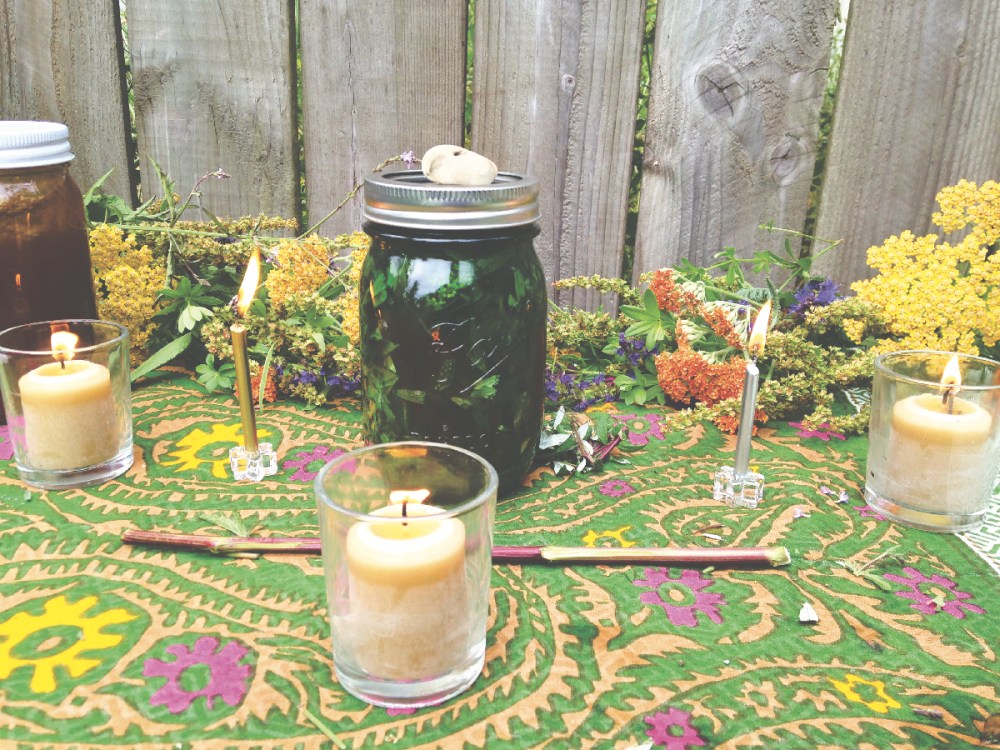 a table covered with herbs, candles, liquor, honey, stones, and a green glass jar