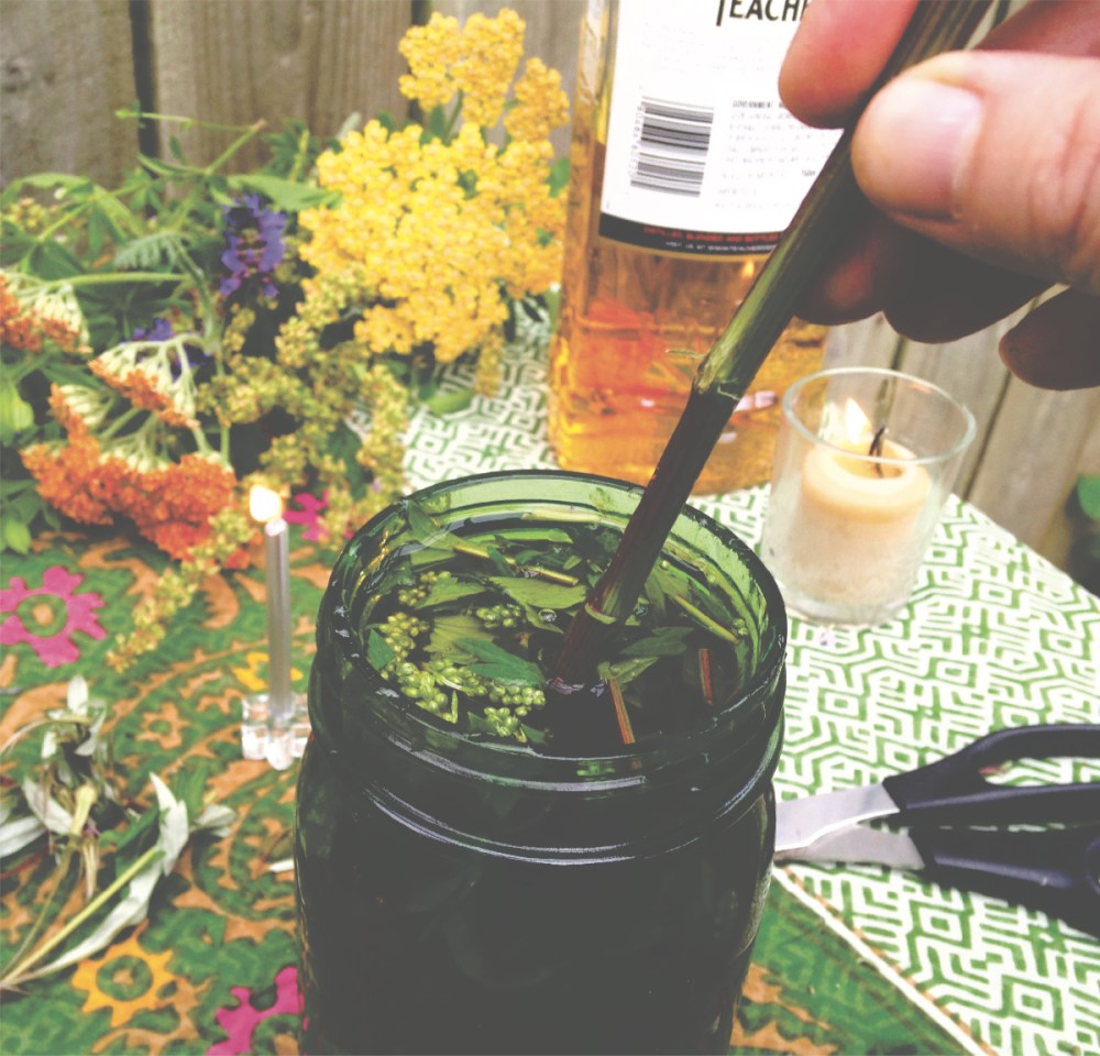 a hand stirring the contents of a jar of liquid and herbs