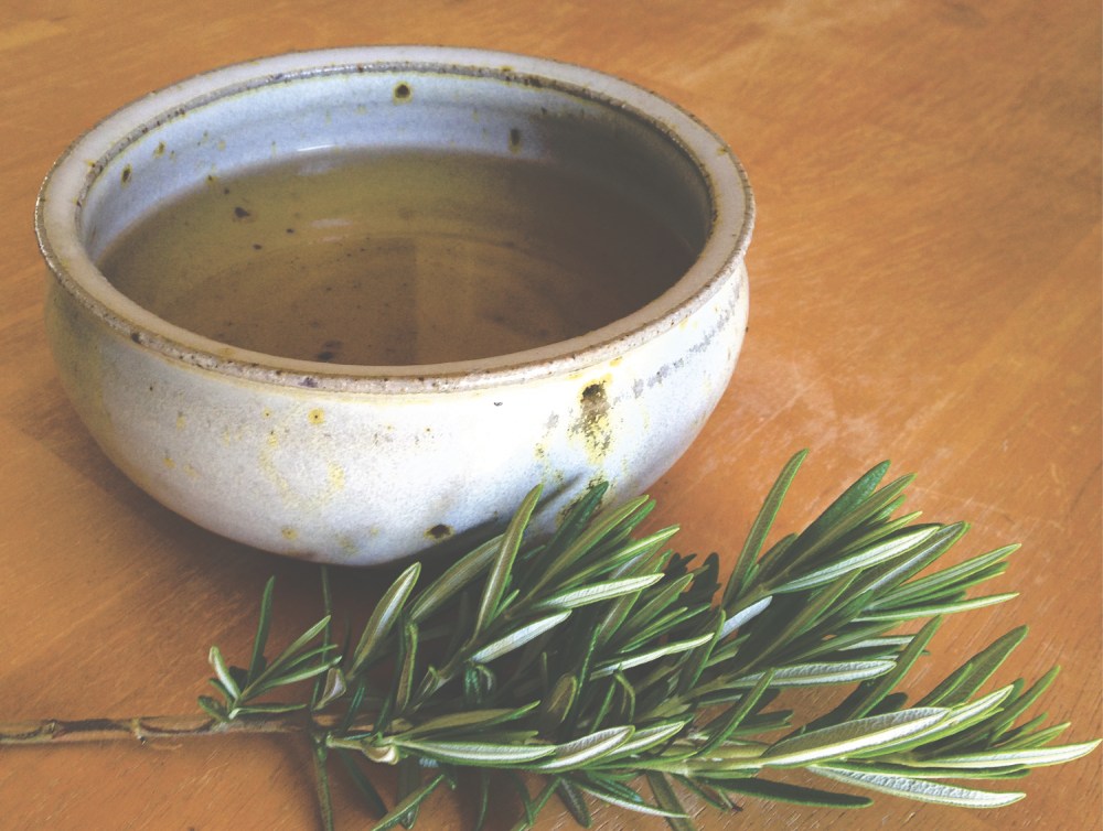 a pottery bowl filled with an herbal infusion beside a fresh sprig of rosemary