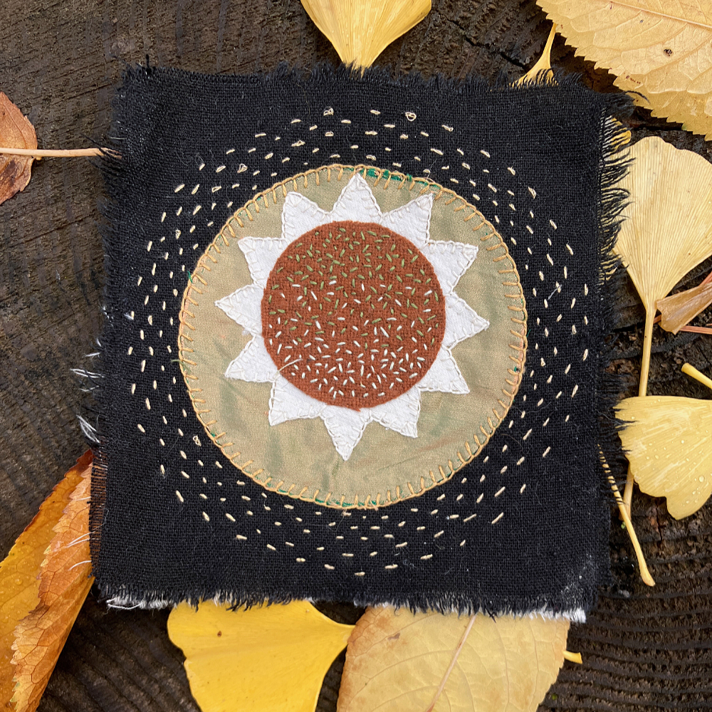 a square of black linen with concentric circles of golden yellow stitching surrounding a circle of golden silk, a 12 pointed star of white silk, and a circle of rust brown wool stitched with dark and light green seed stitch.