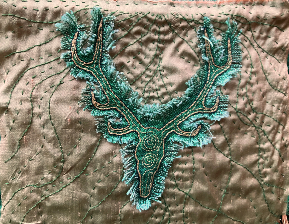 A closeup of green woven fabric embroidered with an antlered stag head on a gold silk background