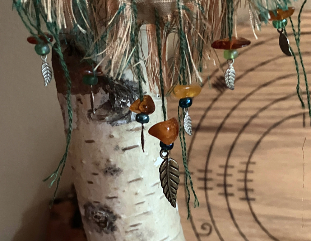 Closeup of amber beads, green glas beads, and brass leaf charms decorating the fringe on the bottom of an embroidered wall hanging