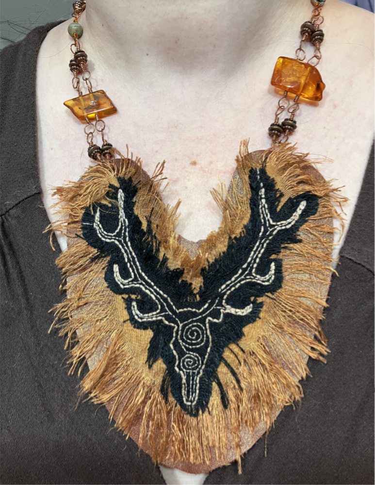 A neclace with a large pendant made of leather and bronze colored silk, adorned with a stag embroidered in cream stitching on black linen. The pendant hangs from copper wire adorned with copper beads and large amber beads.