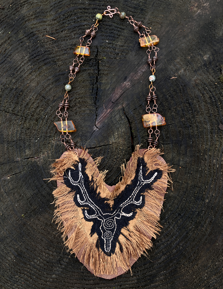 A neclace with a large pendant made of leather and bronze colored silk, adorned with a stag embroidered in cream stitching on black linen. The pendant hangs from copper wire adorned with copper beads and large amber beads.