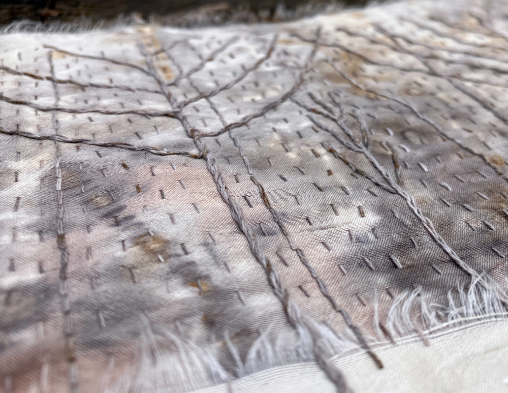 A detail of a stitched art piece featuring white silk, and silvery gray, white, and tan mottled silk, stitched with stylized, bare branched winter trees.