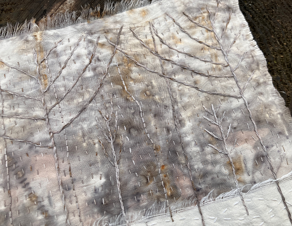 A detail of a stitched art piece featuring white silk, and silvery gray, white, and tan mottled silk, stitched with stylized, bare branched winter trees.