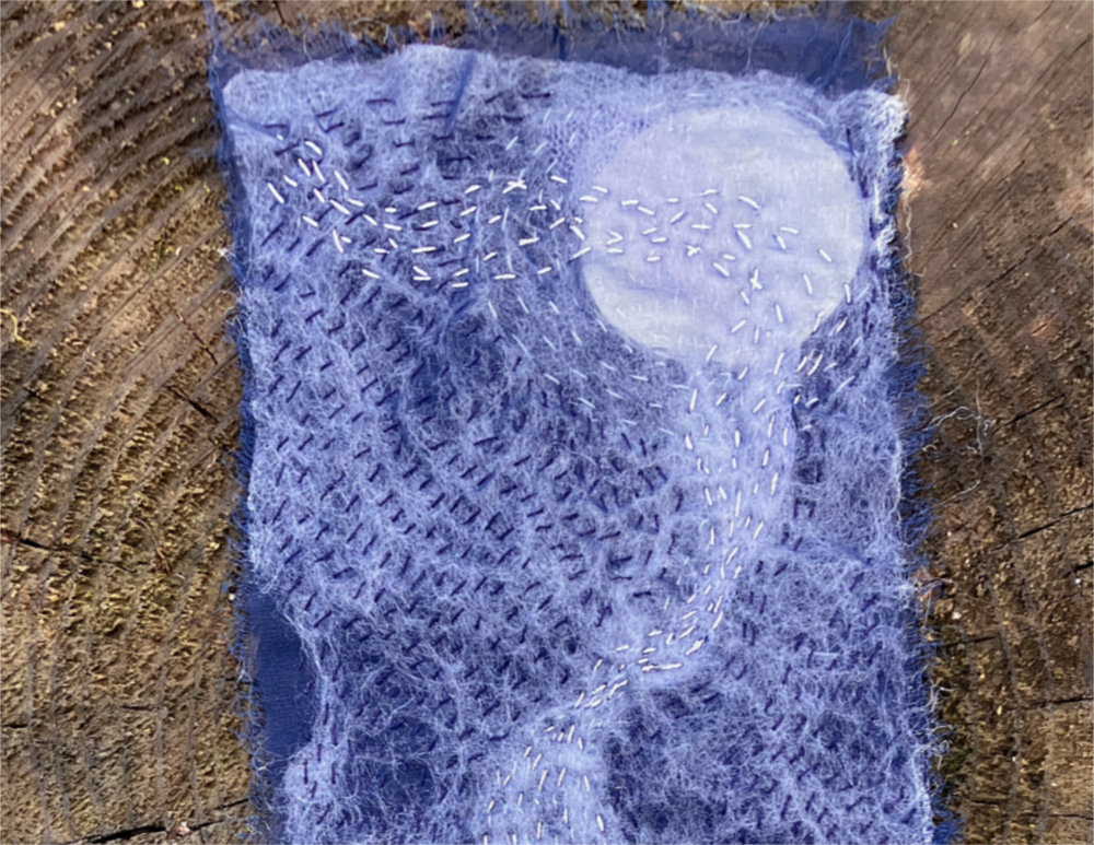 Closeup of blue and white fabric with dark blue, white, and silver stitching, creating a stylized representation of moonlight on moving water