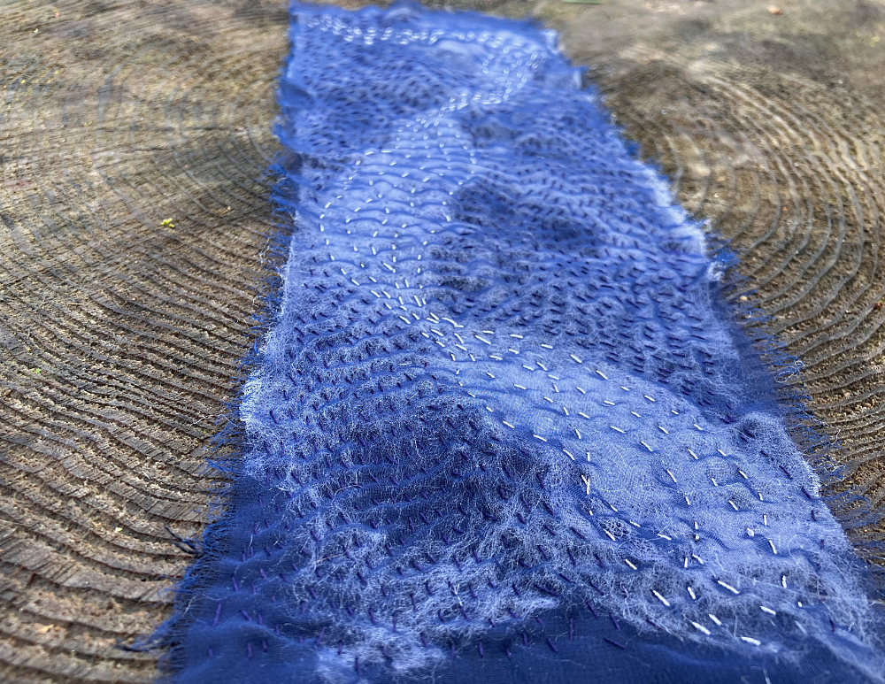 A blue and white rectangle of fabric with dark blue, white, and silver stitching, creating a stylized representation of moonlight on moving water