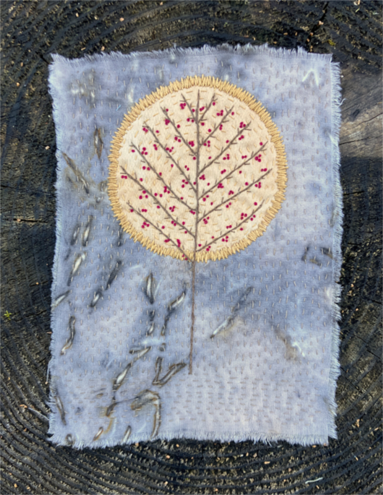 An embroidered, leafless hawthorn tree with red berries, surrounded by a golden silk halo, on a background of fabric in shades of gray.