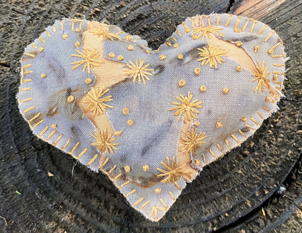A hand stitched heart with gray and rust mottled fabric, with cracks revealing golden yellow fabric underneath, embroidered with small yellow flowers