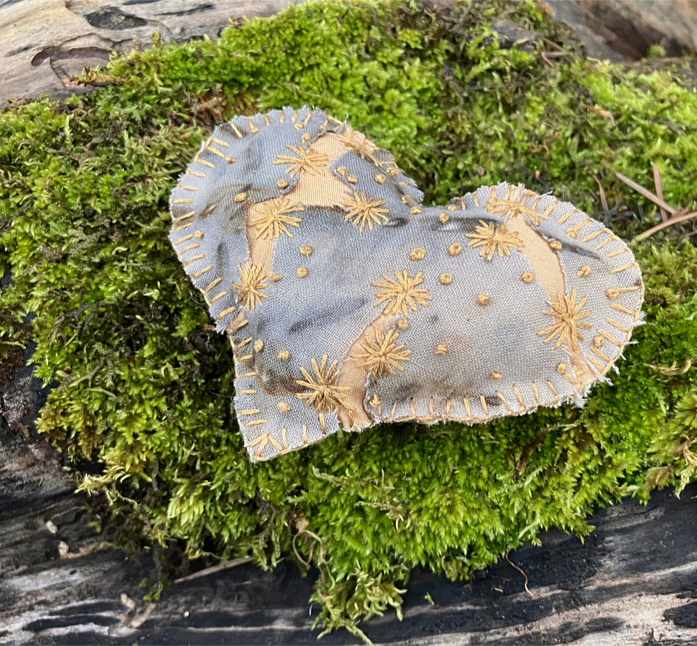 A hand stitched heart with gray and rust mottled fabric, with cracks revealing golden yellow fabric underneath, embroidered with small yellow flowers. The heart sits on a bed of moss.