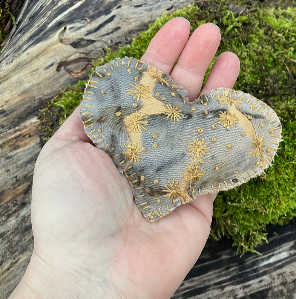 A hand stitched heart with gray and rust mottled fabric, with cracks revealing golden yellow fabric underneath, embroidered with small yellow flowers. The heart is held in a woman's hand.