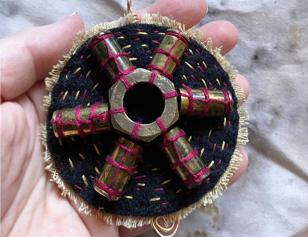 A protective talisman featuring six empty bullet cases arranged around a large brass nut in a radial shape, stitched to a circle of black wool with red thread.