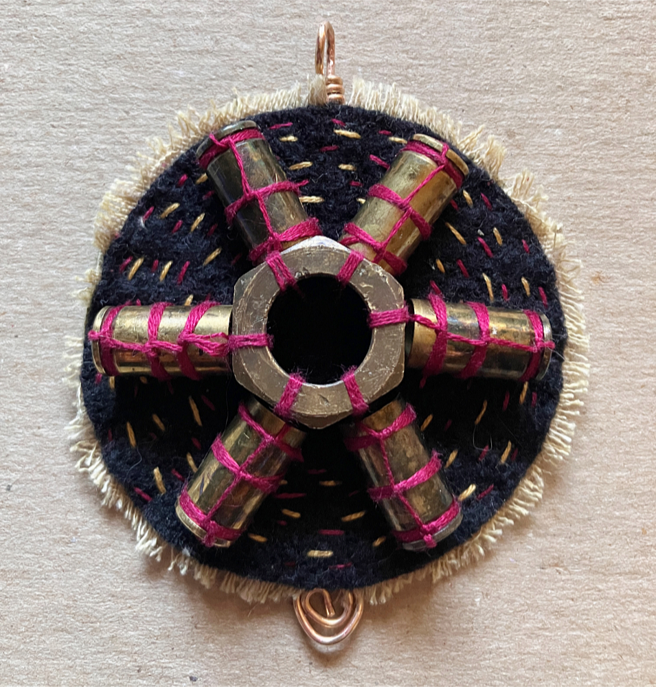 A protective talisman featuring six empty bullet cases arranged around a large brass nut in a radial shape, stitched to a circle of black wool with red thread.
