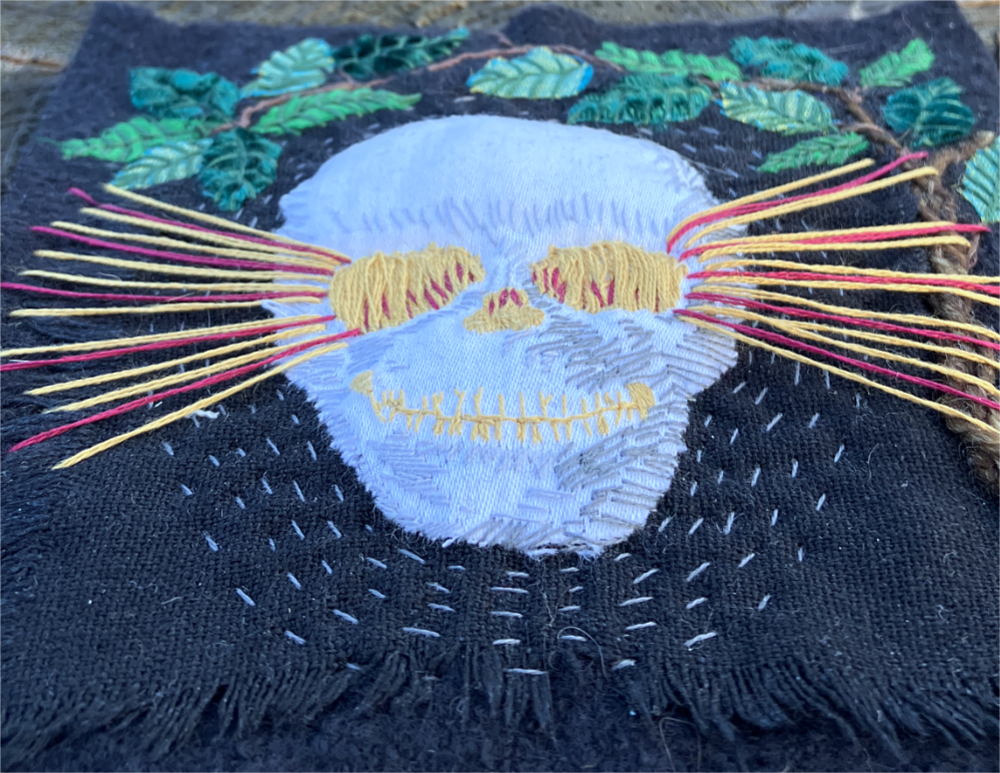 A white skull embroidered on black fabric, surrounded by appliqued leaves, with beams of red and gold light radiating from the eye sockets