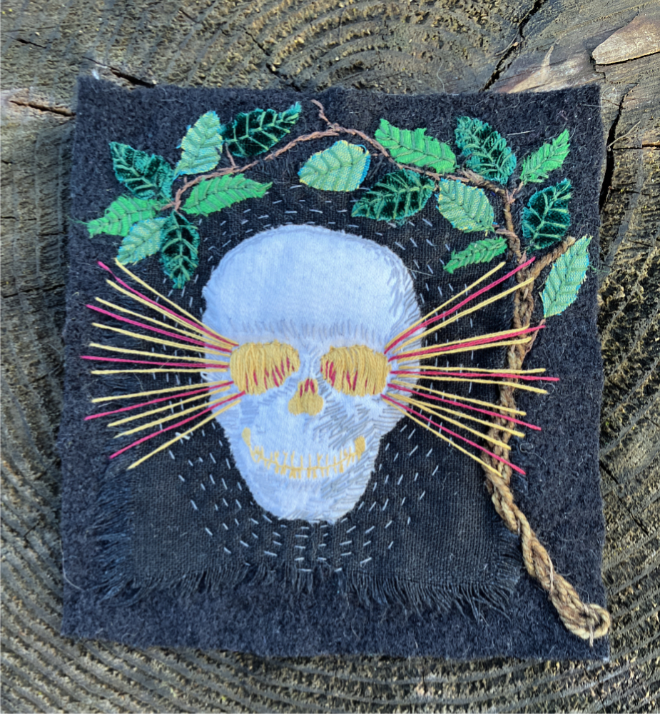 A white skull embroidered on black fabric, surrounded by appliqued leaves, with beams of red and gold light radiating from the eye sockets