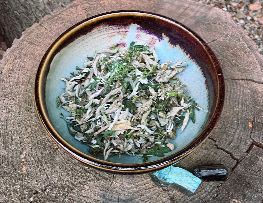 A bowl of dried herbs, with a piece of tourmaline, and a piece of labradorite, sitting next to it on a stump.